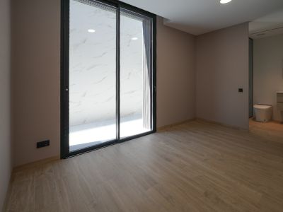 Espaciosa estancia con suelo de gres imitación madera y paredes claras. Grandes puertas correderas de cristal negro dan acceso a una terraza exterior con azulejos de mármol blanco. Baño integrado visible.