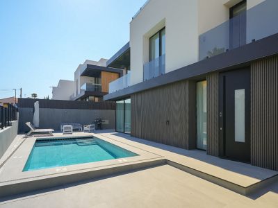 Exterior de villa moderna con piscina rectangular y escaleras integradas, rodeada de pavimento claro. Destaca la fachada inferior con lamas oscuras y la puerta de entrada principal.