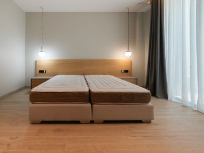 Habitación con cama doble de bases tapizadas, cabecero de madera clara con mesitas de noche integradas y dos lámparas colgantes modernas. Ventanal con cortinas claras.