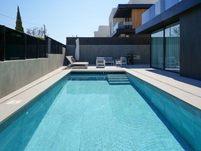 La zona exterior presenta una piscina rectangular con azulejos celestes y escaleras, rodeada de pavimento gris claro. Un moderno conjunto de salón y tumbona invitan a la relajación junto a la elegante fachada.