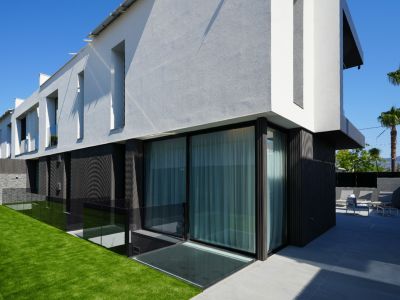 Moderna fachada exterior con contraste de texturas en blanco y gris, grandes ventanales, jardín de césped artificial y patio con mobiliario exterior.