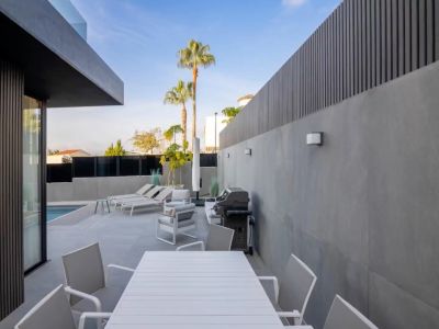 Moderna terraza exterior con mesa de comedor y sillas blancas, zona lounge y barbacoa, enmarcada por paredes de gres gris y celosías oscuras, con vista a la piscina.