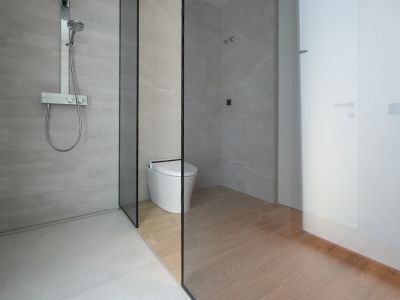 Moderno baño con ducha a ras de suelo, azulejos grises claros y grifería termostática cromada. Una mampara de cristal separa la ducha del inodoro inteligente con suelo de baldosas imitación madera.