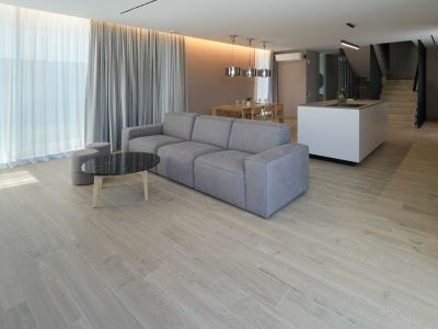 Sala de estar diáfana con sofá modular gris, mesa de centro de mármol negro y suelo de gres porcelánico. Grandes ventanales con cortinas dobles.