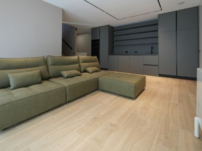 Sofá modular verde oliva en sala de estar de concepto abierto con suelo de baldosas imitación madera. Al fondo, mobiliario de cocina oscuro con fregadero y estantes.