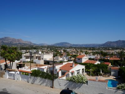 Vista elevada del barrio residencial de Albir, mostrando diversidad arquitectónica, exuberante vegetación y una piscina privada, con la imponente sierra al fondo bajo un cielo despejado.