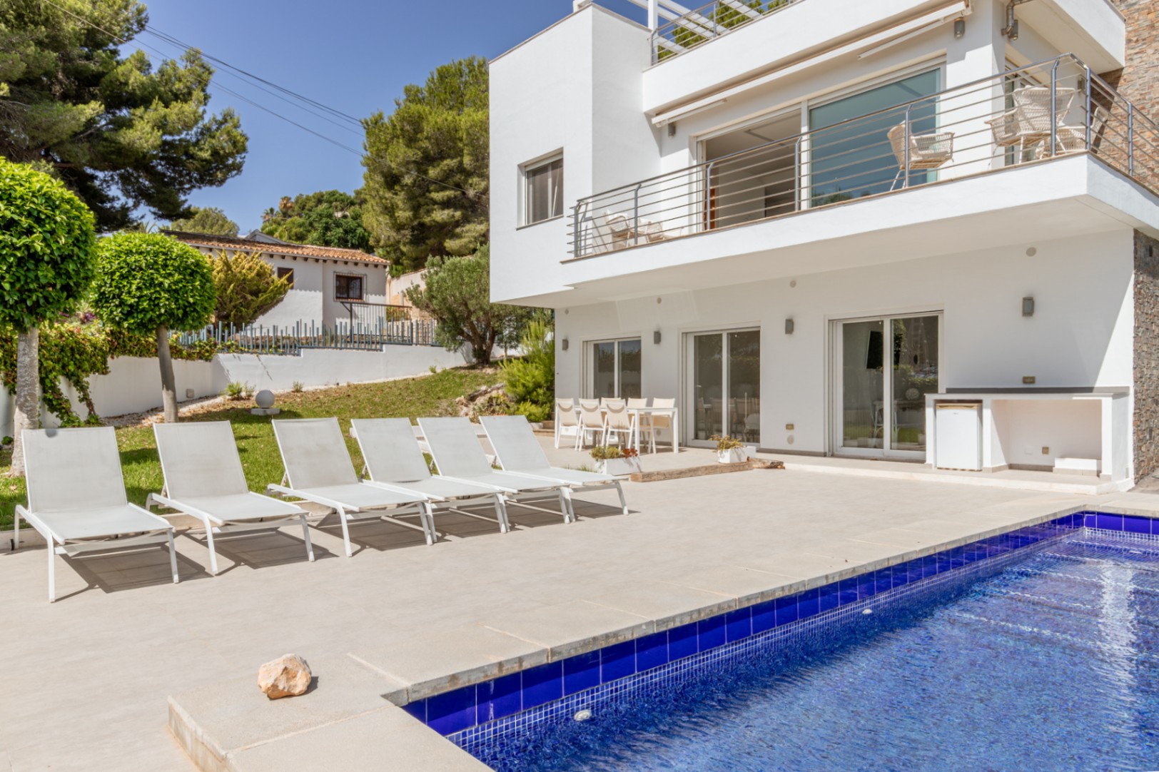 Amplia terraza embaldosada con piscina de mosaico azul, seis tumbonas blancas y comedor exterior. Un área de cocina integrada con nevera complementa este espacio junto a la moderna villa. Amplia terraza embaldosada con piscina de mosaico azul, seis tumbonas blancas y comedor exterior. Un área de cocina integrada con nevera complementa este espacio junto a la moderna villa.