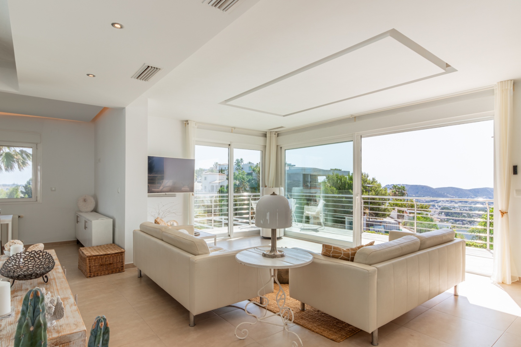 Amplio salón con sofás claros y grandes ventanales correderos que acceden a un balcón. Vistas panorámicas a las colinas y el entorno desde este luminoso espacio con TV mural y luz empotrada. Amplio salón con sofás claros y grandes ventanales correderos que acceden a un balcón. Vistas panorámicas a las colinas y el entorno desde este luminoso espacio con TV mural y luz empotrada.
