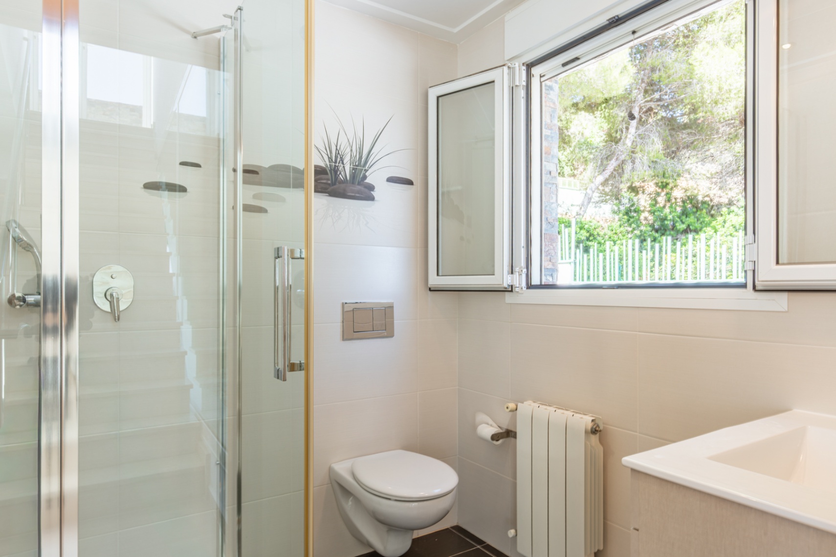 Baño funcional con ducha de cristal, inodoro suspendido y ventana de aluminio blanco que ofrece vistas a la vegetación exterior. Paredes de azulejos beige y radiador. Baño funcional con ducha de cristal, inodoro suspendido y ventana de aluminio blanco que ofrece vistas a la vegetación exterior. Paredes de azulejos beige y radiador.