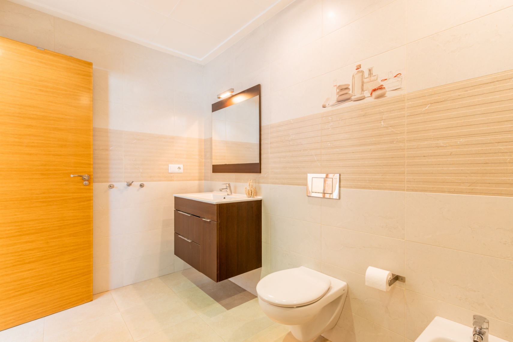 Baño moderno con azulejos claros, puerta de madera, lavabo flotante de madera oscura y espejo. Inodoro suspendido con pulsador cromado y detalles decorativos en la pared. Baño moderno con azulejos claros, puerta de madera, lavabo flotante de madera oscura y espejo. Inodoro suspendido con pulsador cromado y detalles decorativos en la pared.
