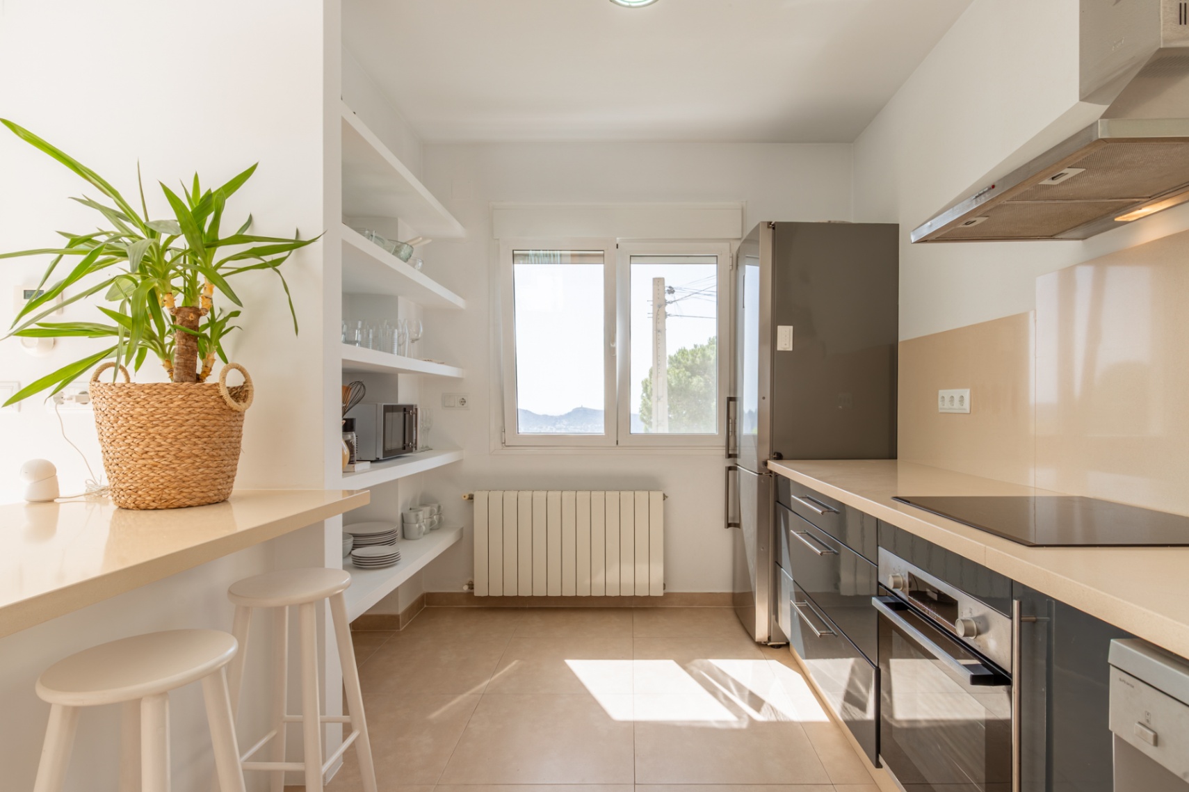 Cocina funcional con muebles oscuros, encimeras claras, placa de inducción, horno integrado y nevera. Barra de desayuno y estantes abiertos bañados por luz natural de la ventana. Cocina funcional con muebles oscuros, encimeras claras, placa de inducción, horno integrado y nevera. Barra de desayuno y estantes abiertos bañados por luz natural de la ventana.