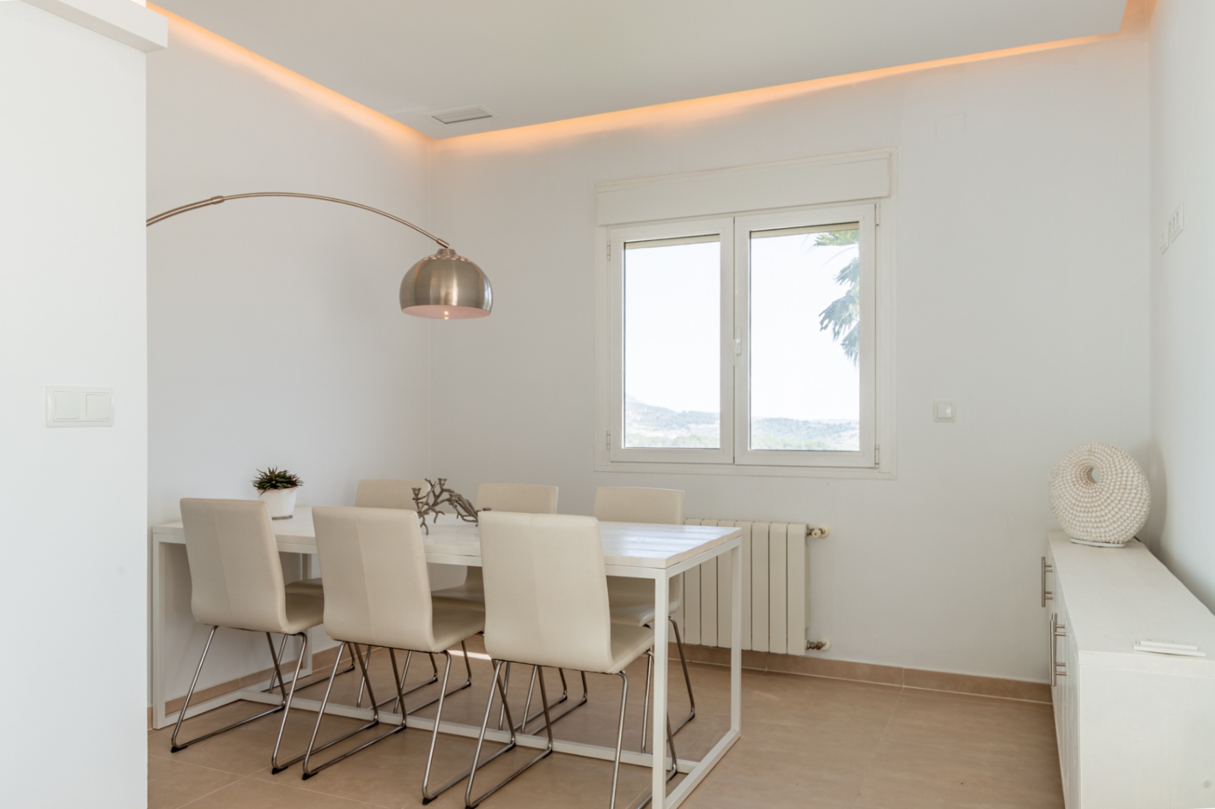 Comedor luminoso con mesa blanca para seis comensales, iluminación ambiental en el techo y vistas a la naturaleza a través de amplios ventanales. Incluye radiador y mobiliario auxiliar. Comedor luminoso con mesa blanca para seis comensales, iluminación ambiental en el techo y vistas a la naturaleza a través de amplios ventanales. Incluye radiador y mobiliario auxiliar.