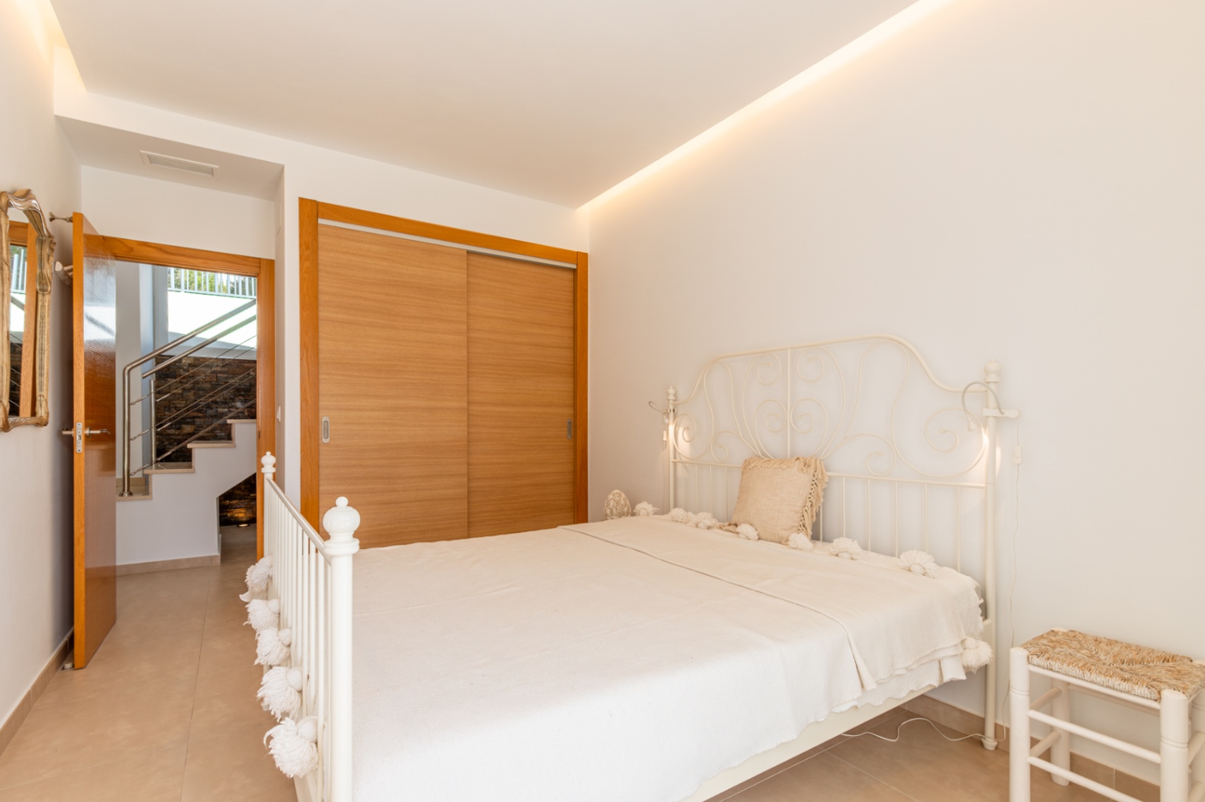Dormitorio luminoso con cama de forja blanca, armario empotrado de madera clara y acceso a escalera interior. Iluminación LED indirecta en el techo. Dormitorio luminoso con cama de forja blanca, armario empotrado de madera clara y acceso a escalera interior. Iluminación LED indirecta en el techo.