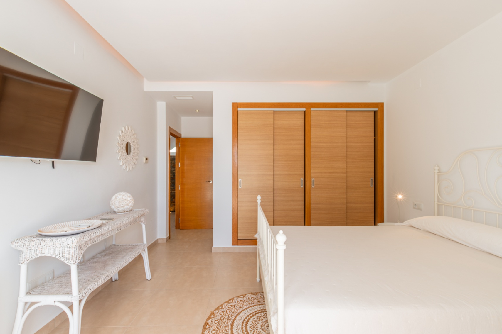 Dormitorio luminoso con cama de forja blanca, armarios empotrados de madera clara y televisor en pared. El espacio incluye una consola de mimbre y suelo de baldosas. Dormitorio luminoso con cama de forja blanca, armarios empotrados de madera clara y televisor en pared. El espacio incluye una consola de mimbre y suelo de baldosas.