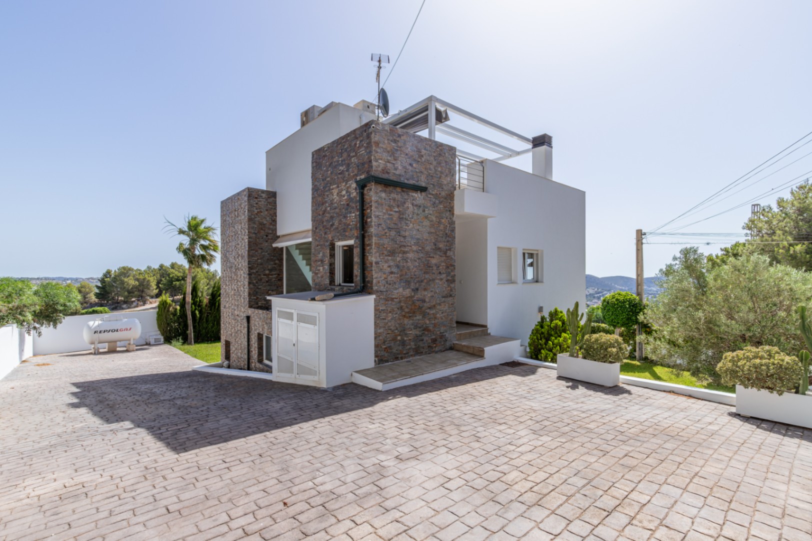 El exterior de la villa moderna presenta revestimiento de piedra, un amplio camino pavimentado y una rampa de acceso. Se observa un tanque de gas Repsol y jardines mediterráneos. El exterior de la villa moderna presenta revestimiento de piedra, un amplio camino pavimentado y una rampa de acceso. Se observa un tanque de gas Repsol y jardines mediterráneos.