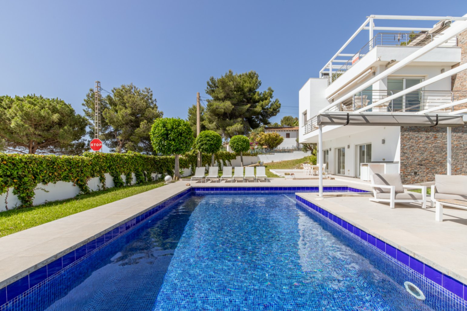 La piscina de 9x4 metros con mosaico azul está rodeada por una terraza solárium de azulejos claros con tumbonas, junto a la moderna villa blanca con revestimiento de piedra y terraza cubierta. La piscina de 9x4 metros con mosaico azul está rodeada por una terraza solárium de azulejos claros con tumbonas, junto a la moderna villa blanca con revestimiento de piedra y terraza cubierta.