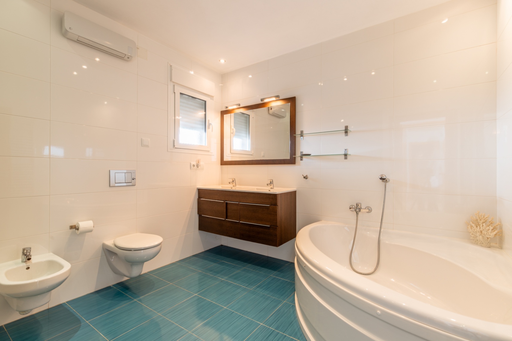 Luminoso baño con bañera de esquina, doble lavabo de madera oscura y inodoro suspendido. Azulejos blancos contrastan con un vibrante suelo turquesa, con luz natural y aire acondicionado. Luminoso baño con bañera de esquina, doble lavabo de madera oscura y inodoro suspendido. Azulejos blancos contrastan con un vibrante suelo turquesa, con luz natural y aire acondicionado.
