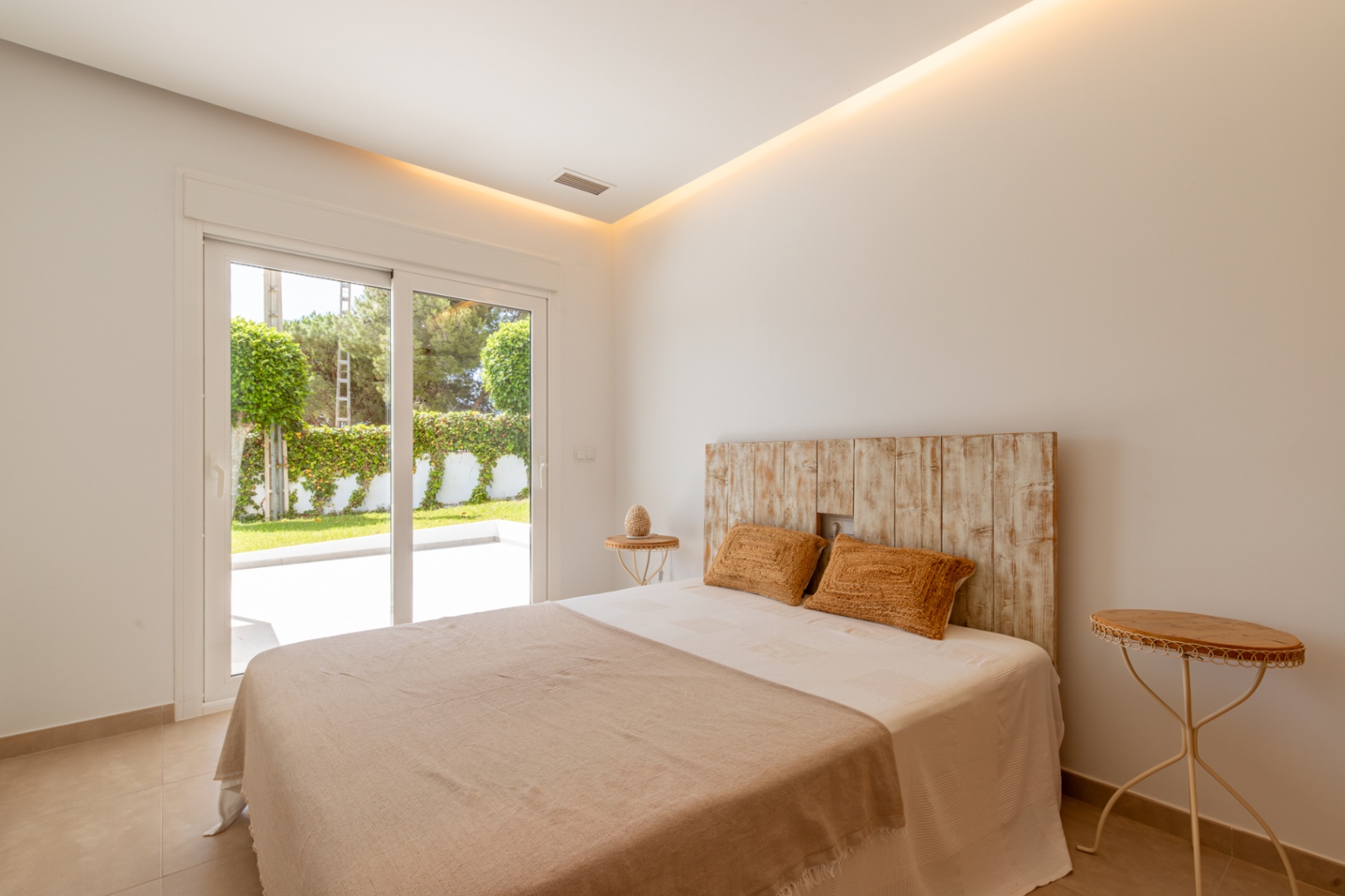 Luminoso dormitorio con cabecero de madera rústica, iluminación de techo integrada y acceso directo a una terraza ajardinada privada mediante puertas correderas. Luminoso dormitorio con cabecero de madera rústica, iluminación de techo integrada y acceso directo a una terraza ajardinada privada mediante puertas correderas.