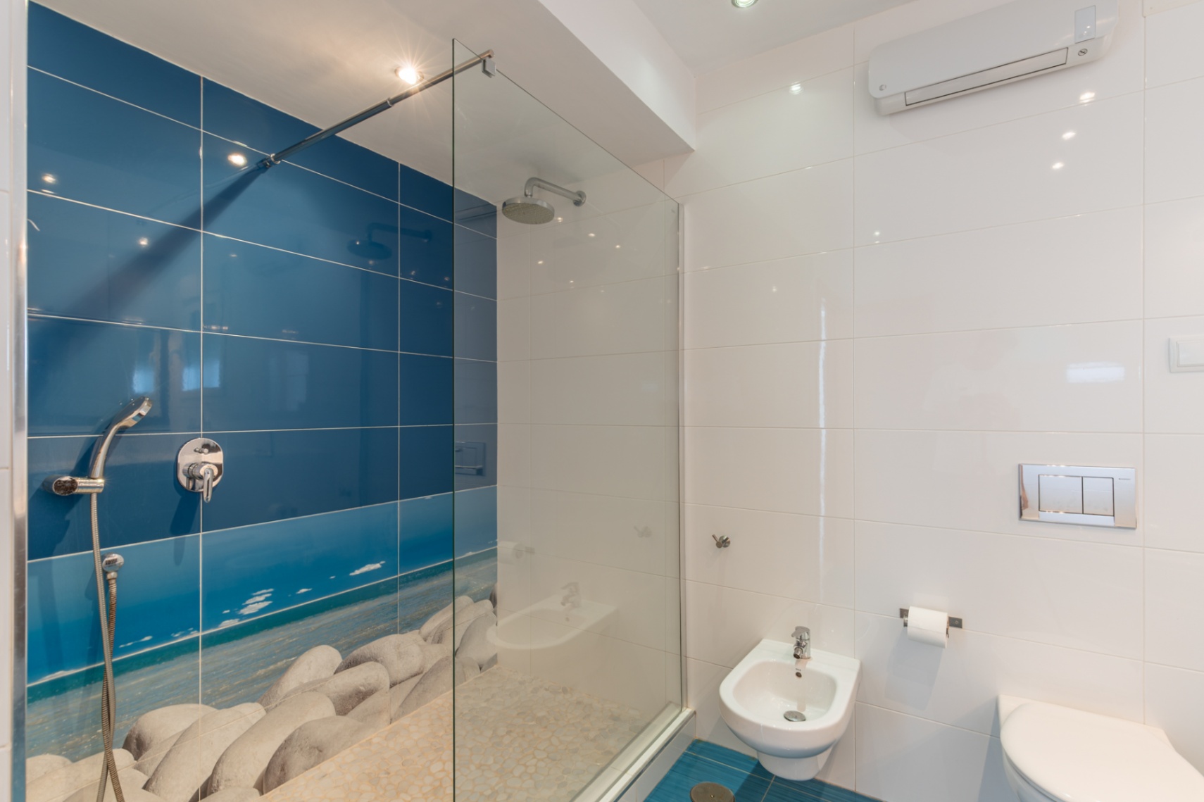 Moderno baño con ducha a ras de suelo y mampara de cristal, pared alicatada en azul con diseño de playa de guijarros y suelo de piedra. Incluye bidé y WC suspendido. Moderno baño con ducha a ras de suelo y mampara de cristal, pared alicatada en azul con diseño de playa de guijarros y suelo de piedra. Incluye bidé y WC suspendido.