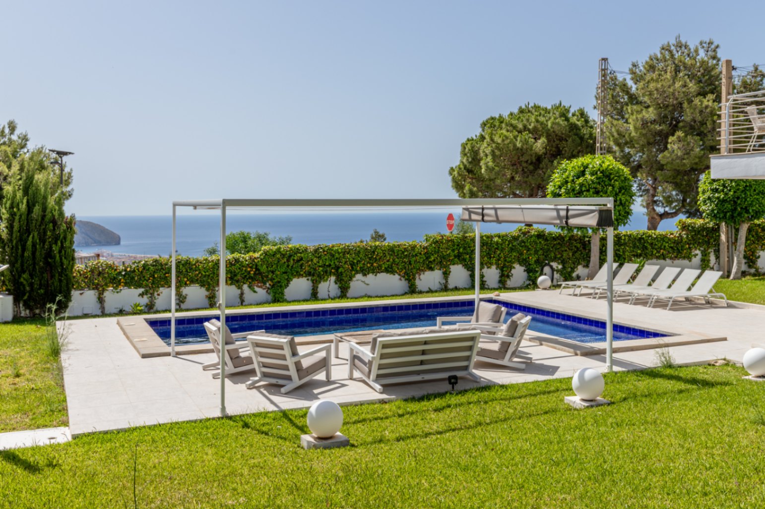 Piscina con azulejos azules y terraza pavimentada con mobiliario lounge bajo una pérgola blanca. Césped cuidado enmarca las vistas abiertas al mar. Piscina con azulejos azules y terraza pavimentada con mobiliario lounge bajo una pérgola blanca. Césped cuidado enmarca las vistas abiertas al mar.
