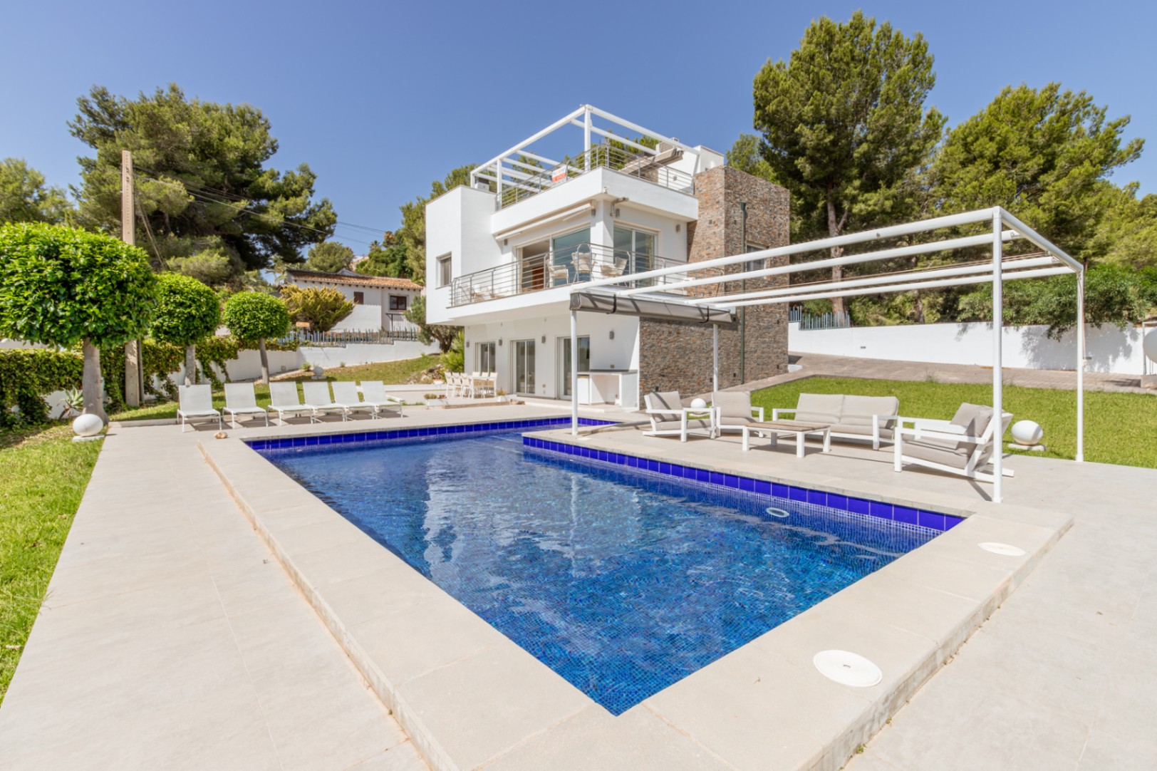 Piscina de mosaico azul y terraza pavimentada con zona chill-out bajo pérgola, complementando la moderna villa con fachada de piedra y balcones. Piscina de mosaico azul y terraza pavimentada con zona chill-out bajo pérgola, complementando la moderna villa con fachada de piedra y balcones.