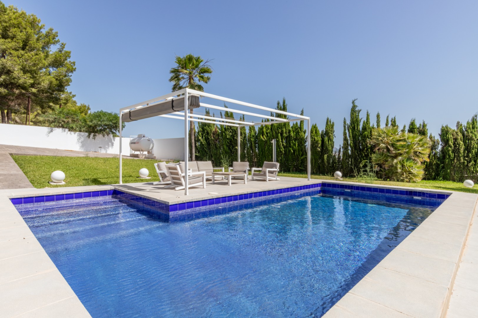 Piscina rectangular de mosaico azul con escaleras, junto a una pérgola blanca moderna con zona lounge y césped bien cuidado. Piscina rectangular de mosaico azul con escaleras, junto a una pérgola blanca moderna con zona lounge y césped bien cuidado.