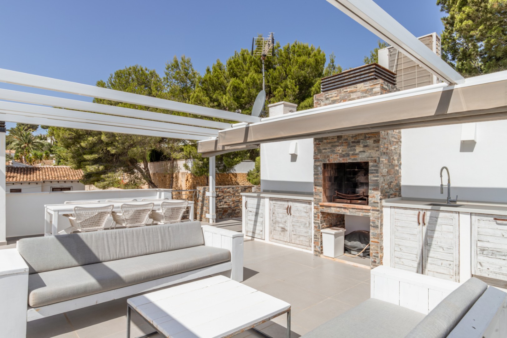 Solárium en azotea con cocina de verano revestida de piedra, barbacoa integrada y fregadero, junto a zonas chill-out y comedor bajo pérgola blanca, rodeado de vegetación. Solárium en azotea con cocina de verano revestida de piedra, barbacoa integrada y fregadero, junto a zonas chill-out y comedor bajo pérgola blanca, rodeado de vegetación.