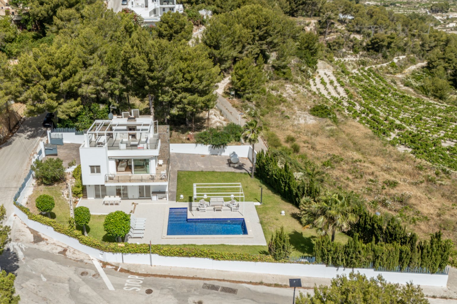 Vista aérea de la villa blanca de tres niveles, con piscina, césped impecable y solárium en la azotea con pérgola. Rodeada de pinos y viñedos. Vista aérea de la villa blanca de tres niveles, con piscina, césped impecable y solárium en la azotea con pérgola. Rodeada de pinos y viñedos.
