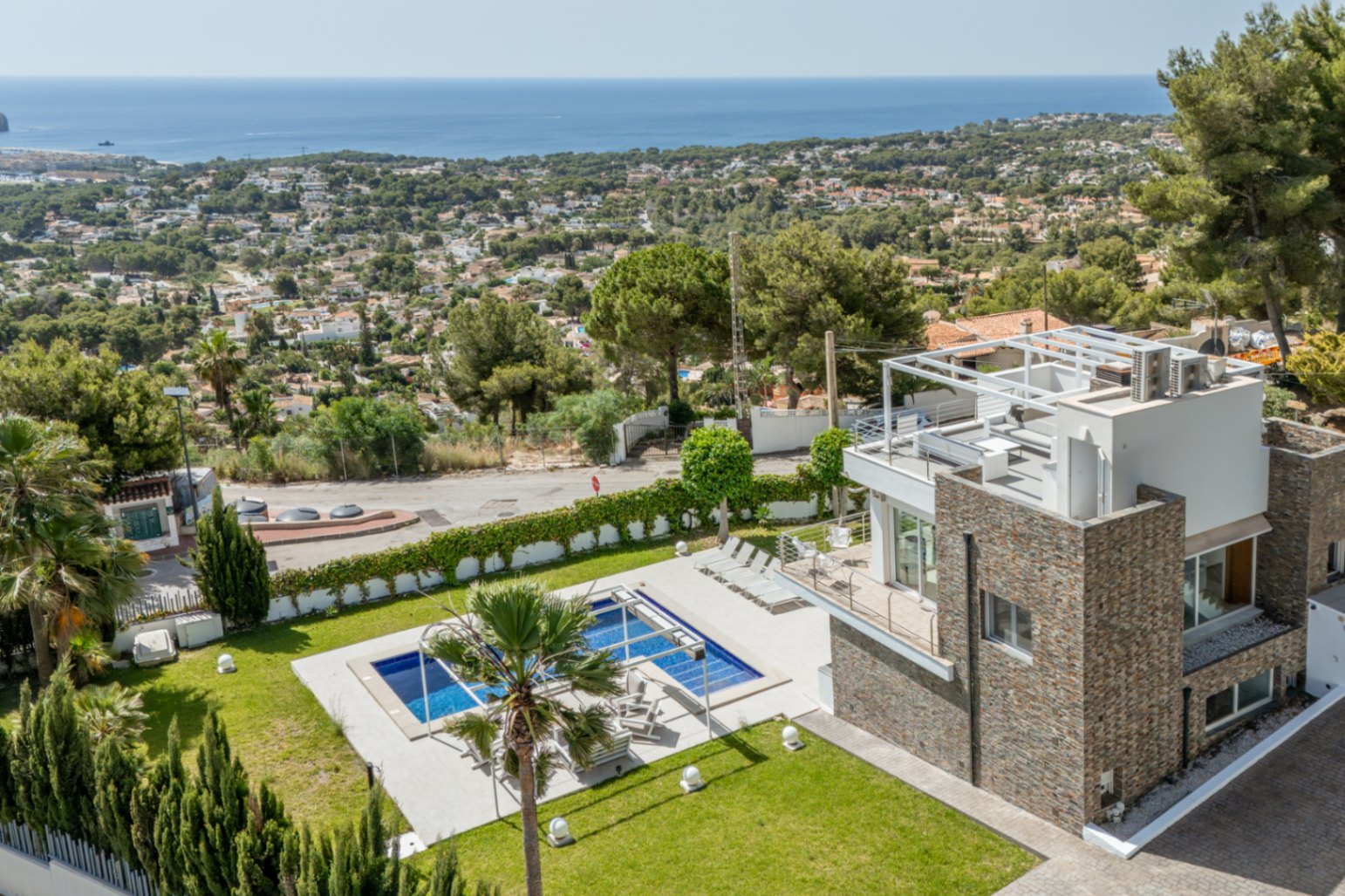 Vista aérea de la villa moderna con fachada de piedra, piscina rectangular, césped cuidado y solárium en la azotea con pérgola, todo con vistas panorámicas al mar. Vista aérea de la villa moderna con fachada de piedra, piscina rectangular, césped cuidado y solárium en la azotea con pérgola, todo con vistas panorámicas al mar.