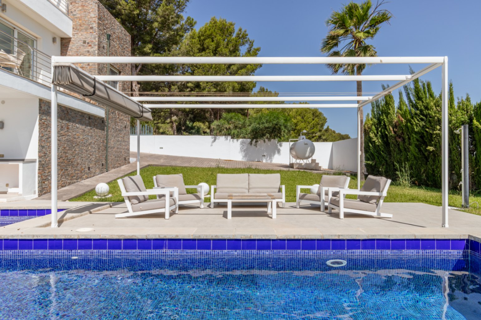 Zona de estar exterior moderna con pérgola blanca y toldo retráctil, situada en una terraza pavimentada junto a la piscina de mosaico azul y jardines. Zona de estar exterior moderna con pérgola blanca y toldo retráctil, situada en una terraza pavimentada junto a la piscina de mosaico azul y jardines.
