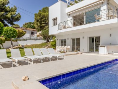 Amplia terraza embaldosada con piscina de mosaico azul, seis tumbonas blancas y comedor exterior. Un área de cocina integrada con nevera complementa este espacio junto a la moderna villa.