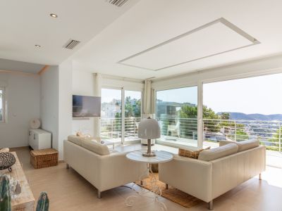 Amplio salón con sofás claros y grandes ventanales correderos que acceden a un balcón. Vistas panorámicas a las colinas y el entorno desde este luminoso espacio con TV mural y luz empotrada.