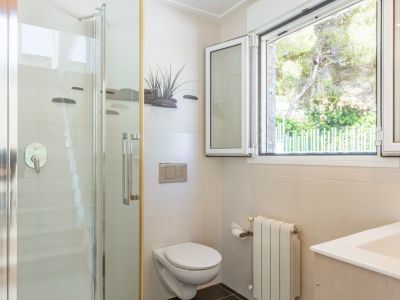 Baño funcional con ducha de cristal, inodoro suspendido y ventana de aluminio blanco que ofrece vistas a la vegetación exterior. Paredes de azulejos beige y radiador.