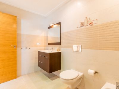 Baño moderno con azulejos claros, puerta de madera, lavabo flotante de madera oscura y espejo. Inodoro suspendido con pulsador cromado y detalles decorativos en la pared.