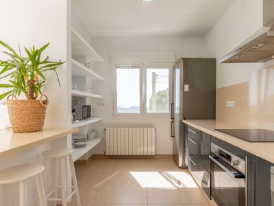 Cocina funcional con muebles oscuros, encimeras claras, placa de inducción, horno integrado y nevera. Barra de desayuno y estantes abiertos bañados por luz natural de la ventana.