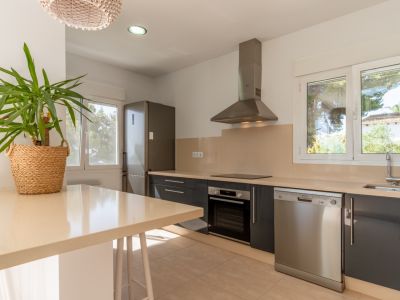 Cocina moderna con mobiliario gris oscuro, encimeras beige claro y electrodomésticos de acero inoxidable. Grandes ventanales aportan luz natural y vistas al exterior.
