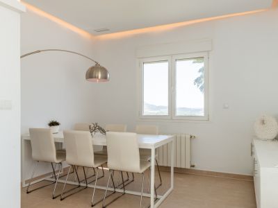 Comedor luminoso con mesa blanca para seis comensales, iluminación ambiental en el techo y vistas a la naturaleza a través de amplios ventanales. Incluye radiador y mobiliario auxiliar.