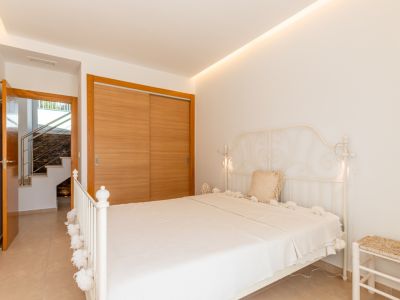Dormitorio luminoso con cama de forja blanca, armario empotrado de madera clara y acceso a escalera interior. Iluminación LED indirecta en el techo.