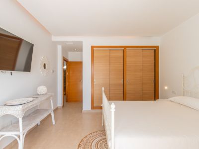 Dormitorio luminoso con cama de forja blanca, armarios empotrados de madera clara y televisor en pared. El espacio incluye una consola de mimbre y suelo de baldosas.