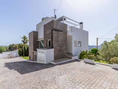 El exterior de la villa moderna presenta revestimiento de piedra, un amplio camino pavimentado y una rampa de acceso. Se observa un tanque de gas Repsol y jardines mediterráneos.