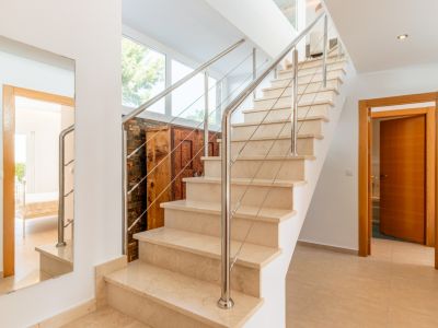 Escalera interior de mármol claro con barandilla de acero inoxidable y cables, iluminada por ventanales que muestran vegetación. Un mueble de madera tallada y un espejo completan el espacio.