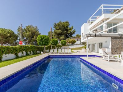 La piscina de 9x4 metros con mosaico azul está rodeada por una terraza solárium de azulejos claros con tumbonas, junto a la moderna villa blanca con revestimiento de piedra y terraza cubierta.