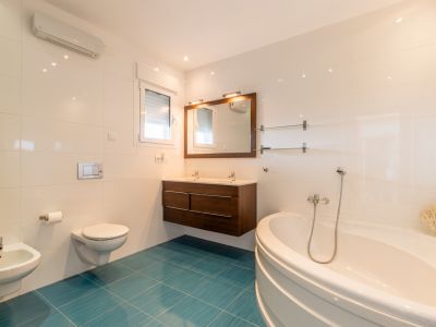 Luminoso baño con bañera de esquina, doble lavabo de madera oscura y inodoro suspendido. Azulejos blancos contrastan con un vibrante suelo turquesa, con luz natural y aire acondicionado.