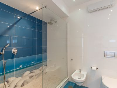 Moderno baño con ducha a ras de suelo y mampara de cristal, pared alicatada en azul con diseño de playa de guijarros y suelo de piedra. Incluye bidé y WC suspendido.