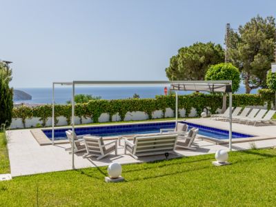 Piscina con azulejos azules y terraza pavimentada con mobiliario lounge bajo una pérgola blanca. Césped cuidado enmarca las vistas abiertas al mar.