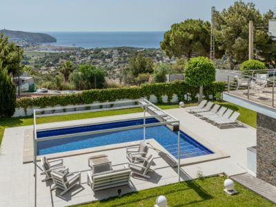 Piscina con cubierta automática y zona chill-out bajo pérgola, en jardín con césped impecable y vistas panorámicas al mar de Moraira.