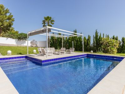 Piscina rectangular de mosaico azul con escaleras, junto a una pérgola blanca moderna con zona lounge y césped bien cuidado.