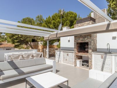 Solárium en azotea con cocina de verano revestida de piedra, barbacoa integrada y fregadero, junto a zonas chill-out y comedor bajo pérgola blanca, rodeado de vegetación.