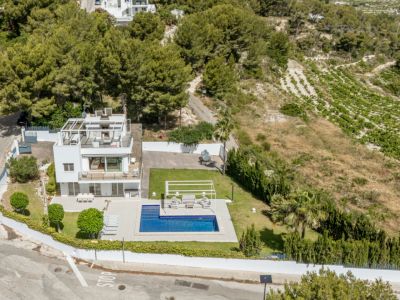 Vista aérea de la villa blanca de tres niveles, con piscina, césped impecable y solárium en la azotea con pérgola. Rodeada de pinos y viñedos.