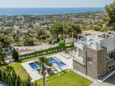 Vista aérea de la villa moderna con fachada de piedra, piscina rectangular, césped cuidado y solárium en la azotea con pérgola, todo con vistas panorámicas al mar.